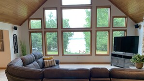 A living room with a sectional sofa, a flat-screen TV, and large windows offering a view of trees. - Beautiful 6 bedroom 3 bath lake front home with view of Lake Nokomis! (Tomahawk)