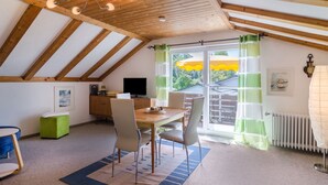 A dining area with a table, chairs, a television, and a view through a window with curtains. - Apartment "Säntisblick" with Beautiful View of Lake Constance and the Alps (Daisendorf)