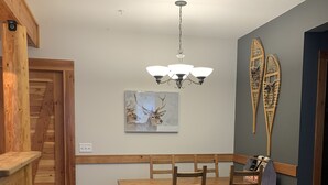 A dining area with a wooden table and chairs, a ceiling light fixture, and a wall-mounted painting of deer. - Big White Condo perfect for an amazing getaway Winter or Summer! (Beaverdell)