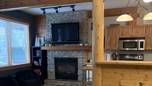 A living room with a stone fireplace, wooden beams, a television, and a kitchen area with a microwave. - Big White Condo perfect for an amazing getaway Winter or Summer! (Beaverdell)