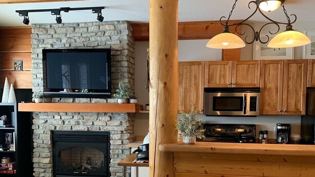 A cozy living area with a stone fireplace, wooden beams, a television, and a kitchen area with modern appliances. - Big White Condo perfect for an amazing getaway Winter or Summer! (Beaverdell)