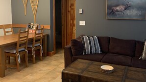 A living room with a brown couch, a wooden coffee table, and a dining area with a wooden table and chairs. - Big White Condo perfect for an amazing getaway Winter or Summer! (Beaverdell)