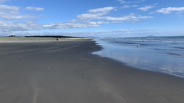 A wide sandy beach with gentle waves, a clear sky, and distant mountains.
