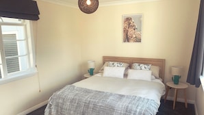 A bedroom with a bed, two bedside tables, a hanging light fixture, a window with blinds, and a framed picture on the wall.