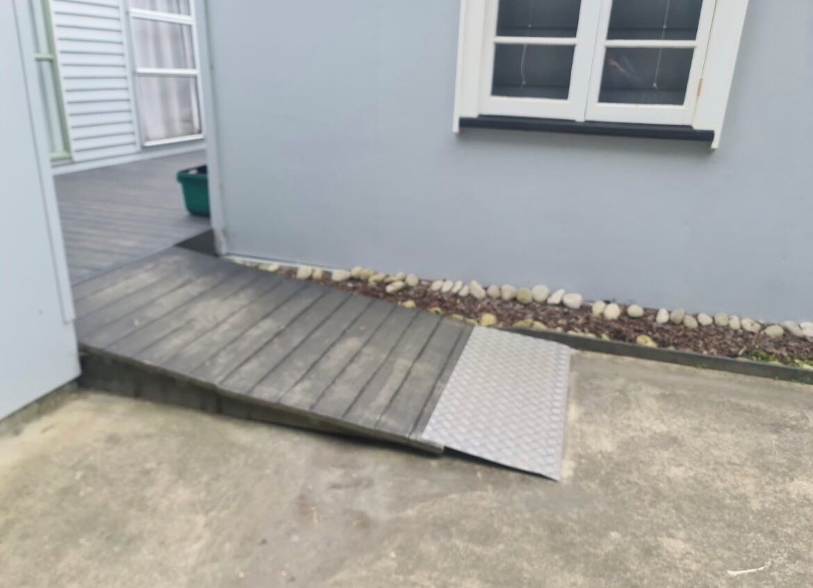 Un patio de concreto con una rampa de madera y una barandilla metálica que conduce a una puerta.