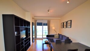 Un salon moderne avec un meuble de rangement noir pour appareils électroniques, un canapé, une table à manger avec des chaises, et une vue sur l’extérieur grâce à une porte vitrée.