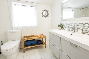 A bathroom with a toilet, a wooden bench with towels, a sink with a mirror, and a tiled backsplash. - Modern House| Walk to the Falls (Niagara Falls)