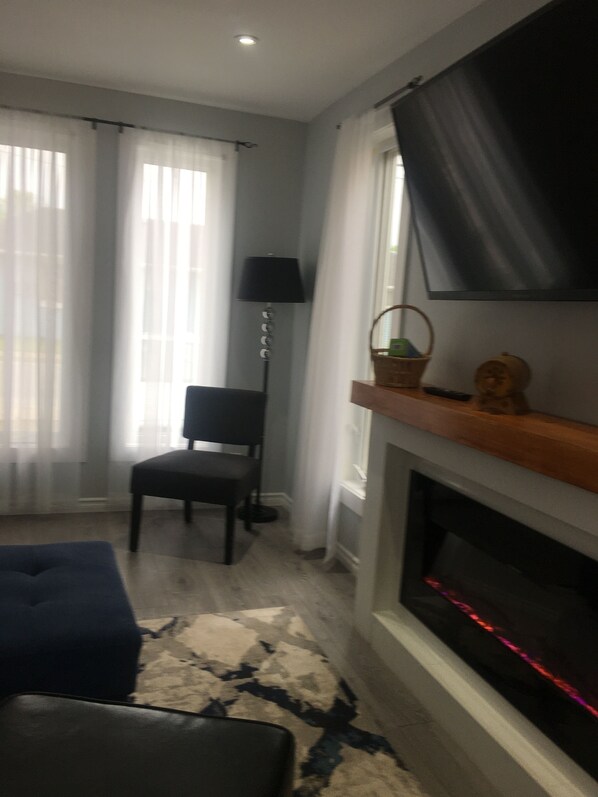 A living room with a fireplace, a television mounted on the wall, a chair, and a sofa. - Modern House| Walk to the Falls (Niagara Falls)