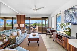 A modern living room with a sofa, coffee table, and TV, featuring large windows and a view of greenery. - Kapalua 1BR Villa Near Beaches and Golf (Lahaina)