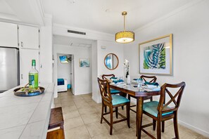 Une cuisine moderne avec des armoires blanches, un coin repas avec des chaises en bois et une table dressée pour deux personnes, ainsi qu’une grande œuvre d’art encadrée accrochée au mur.