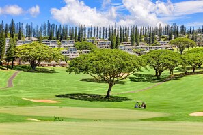 A golf course with a clubhouse, surrounded by a forest of tall trees and a row of residential buildings in the background.