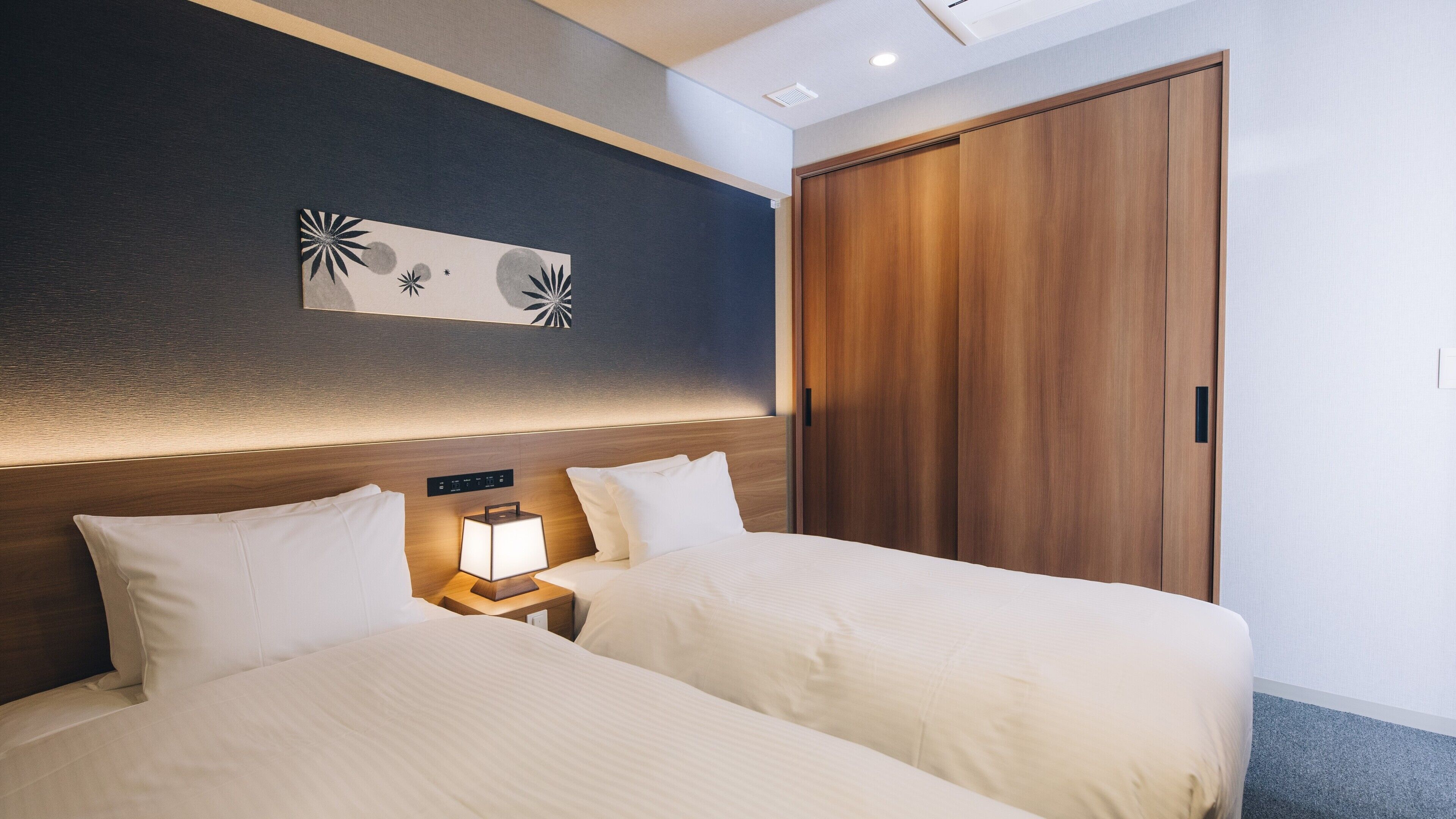 A hotel room with two beds, a nightstand with a lamp, a wooden wardrobe, and a wall with a decorative piece.