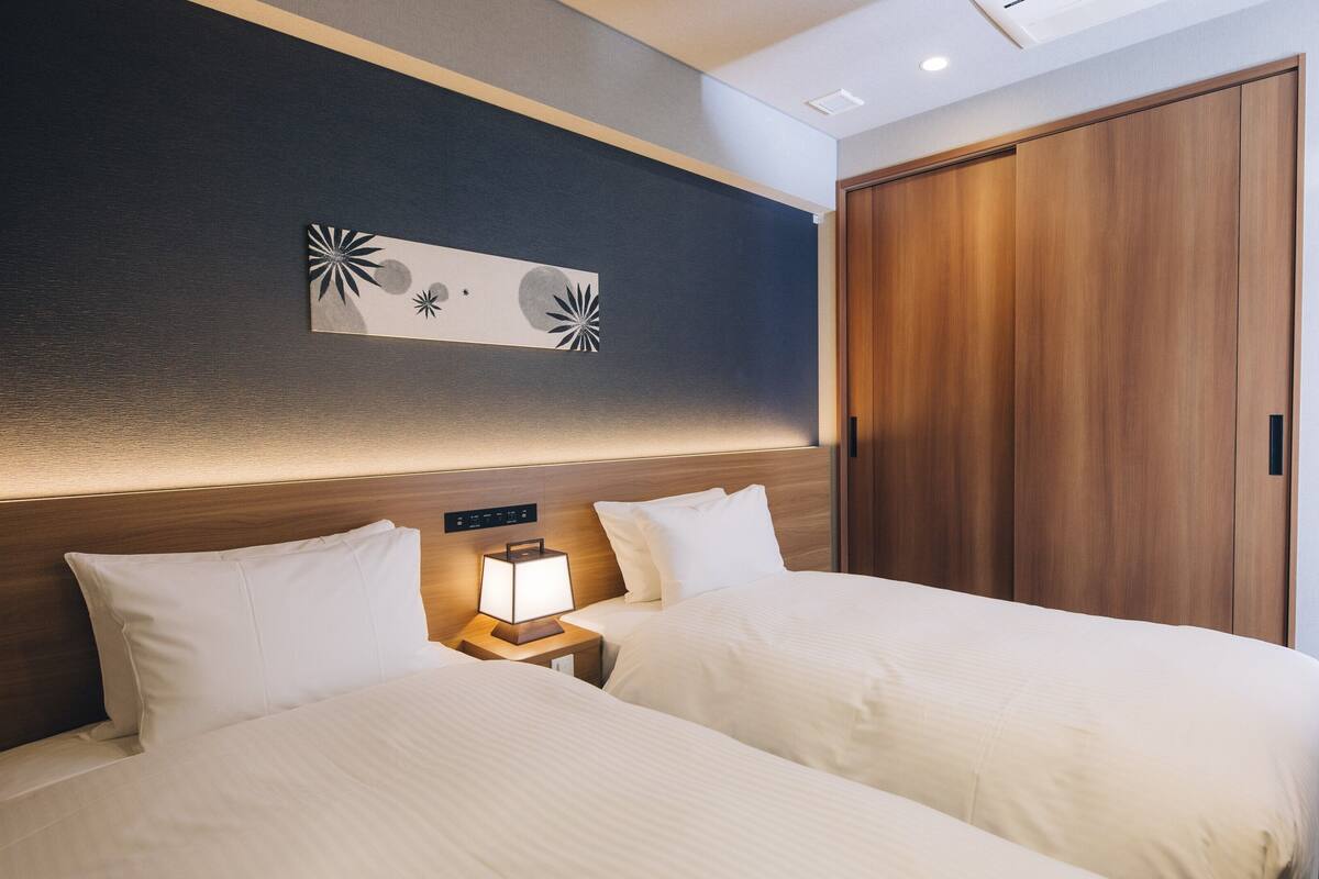 A hotel room with two beds, a nightstand with a lamp, a wooden wardrobe, and a wall with a decorative piece.