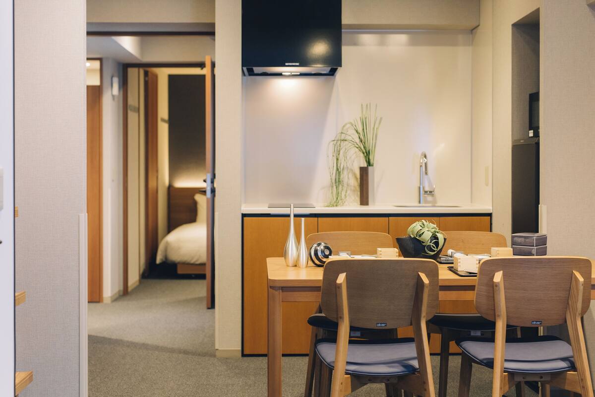 A modern hotel room with a wooden dining table, two chairs, a small potted plant, and a flat-screen TV mounted on the wall.