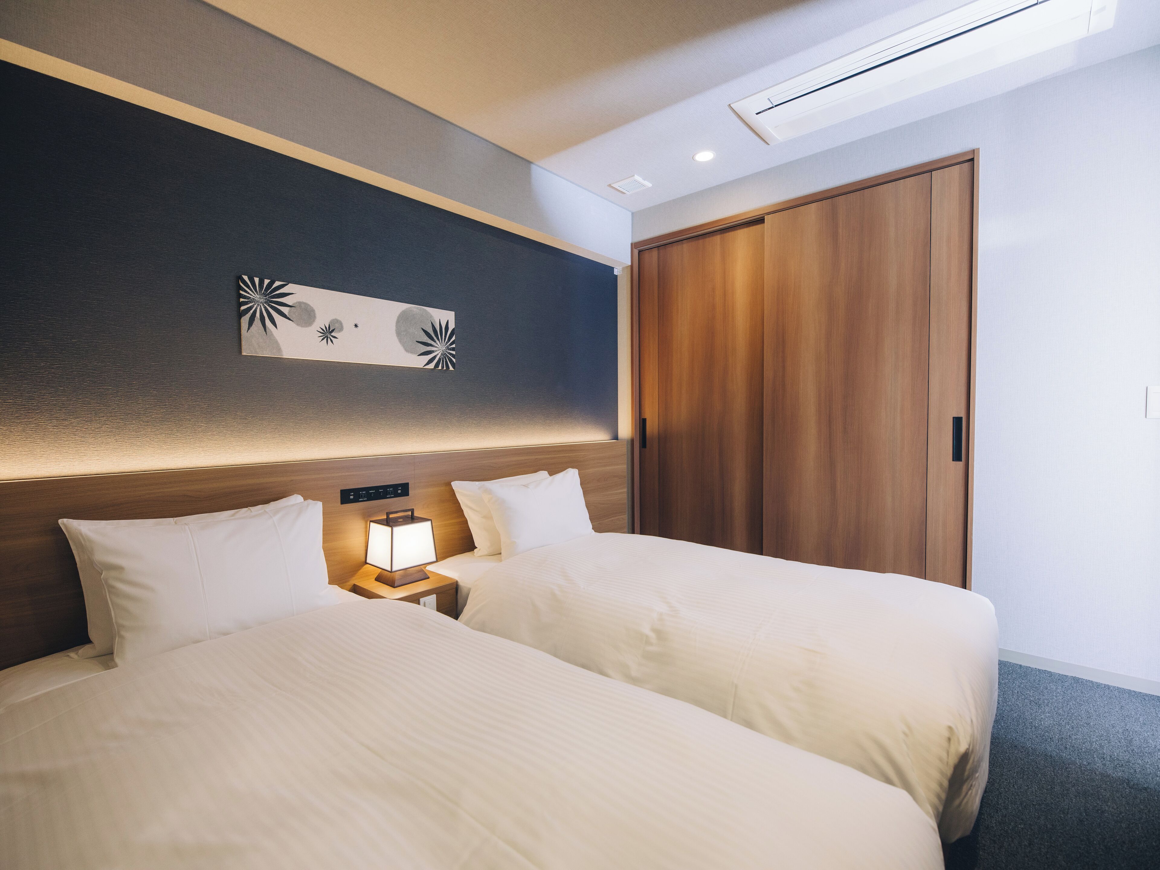 Une chambre d’hôtel avec deux lits, une table de chevet avec une lampe, une armoire en bois et une œuvre d’art accrochée au mur.