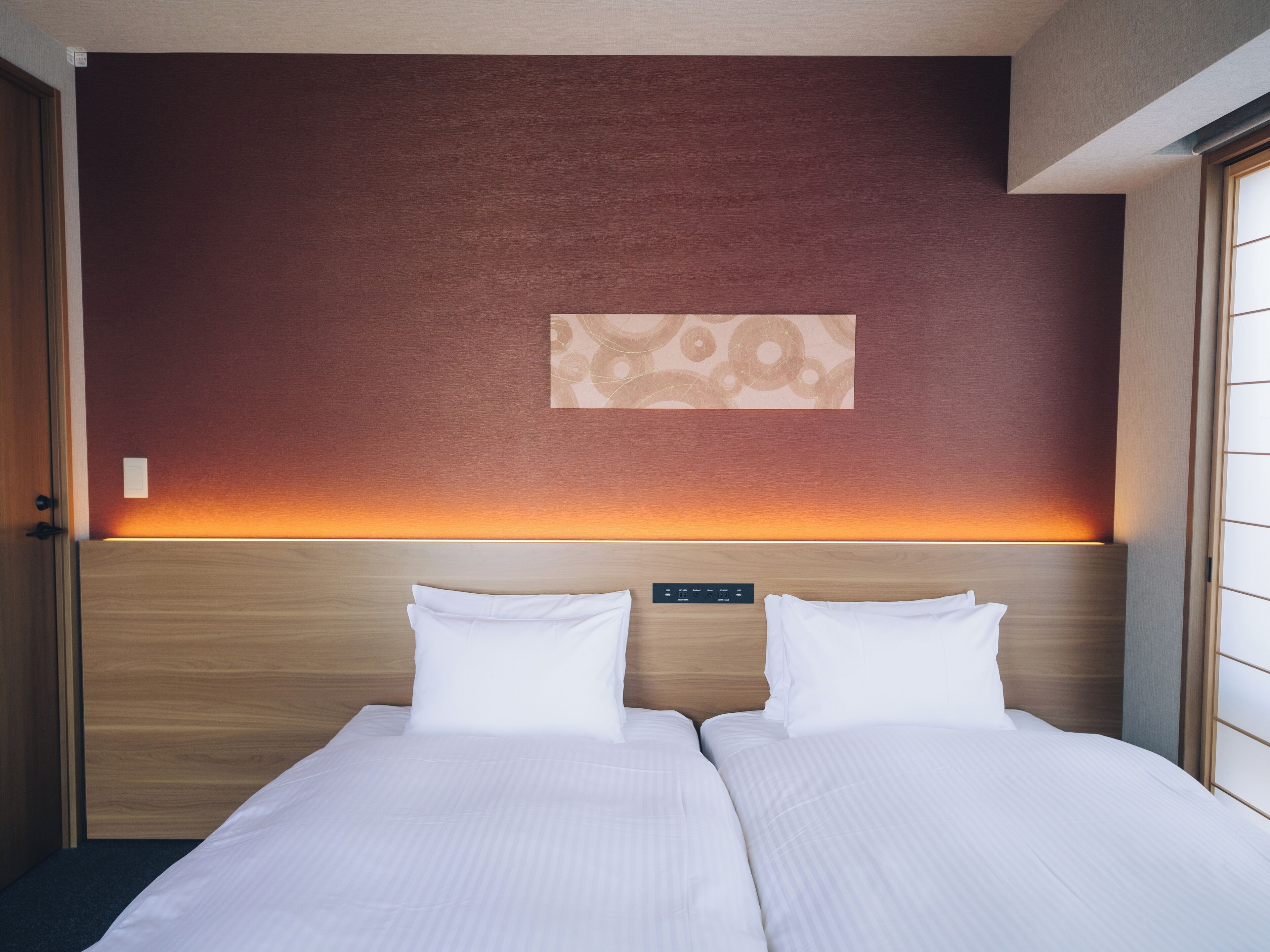 A hotel room with a double bed, a headboard with a control panel, and a wall-mounted artwork.