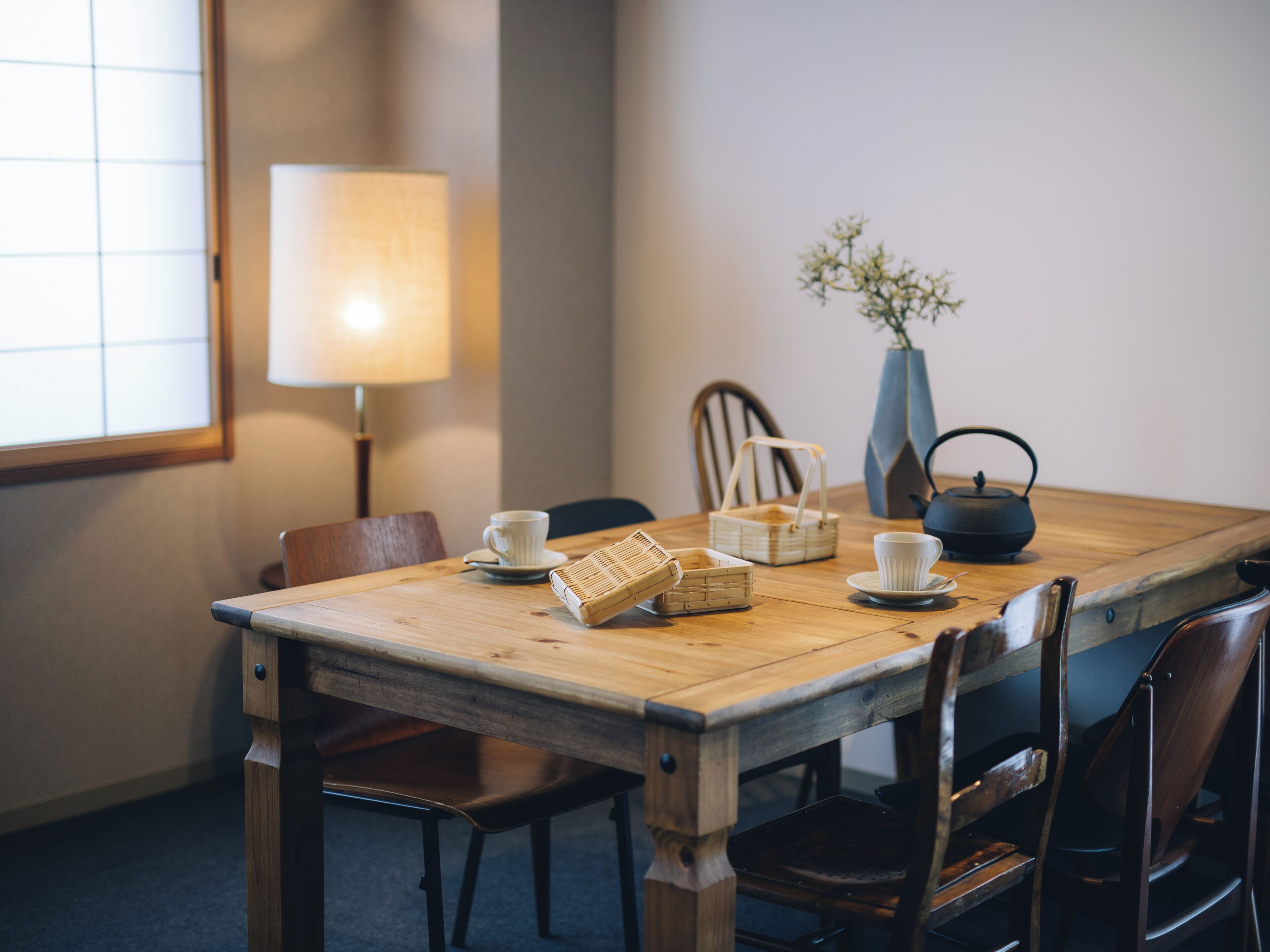 A wooden dining table with chairs, a lamp, a vase with flowers, and a teapot.
