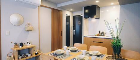 Un rincón comedor compacto con una mesa de madera dispuesta para cuatro personas, rodeada de sillas. A la izquierda, un estante con utensilios de cocina y una cesta tejida. En el fondo, una puerta negra y una pared blanca con un televisor montado.