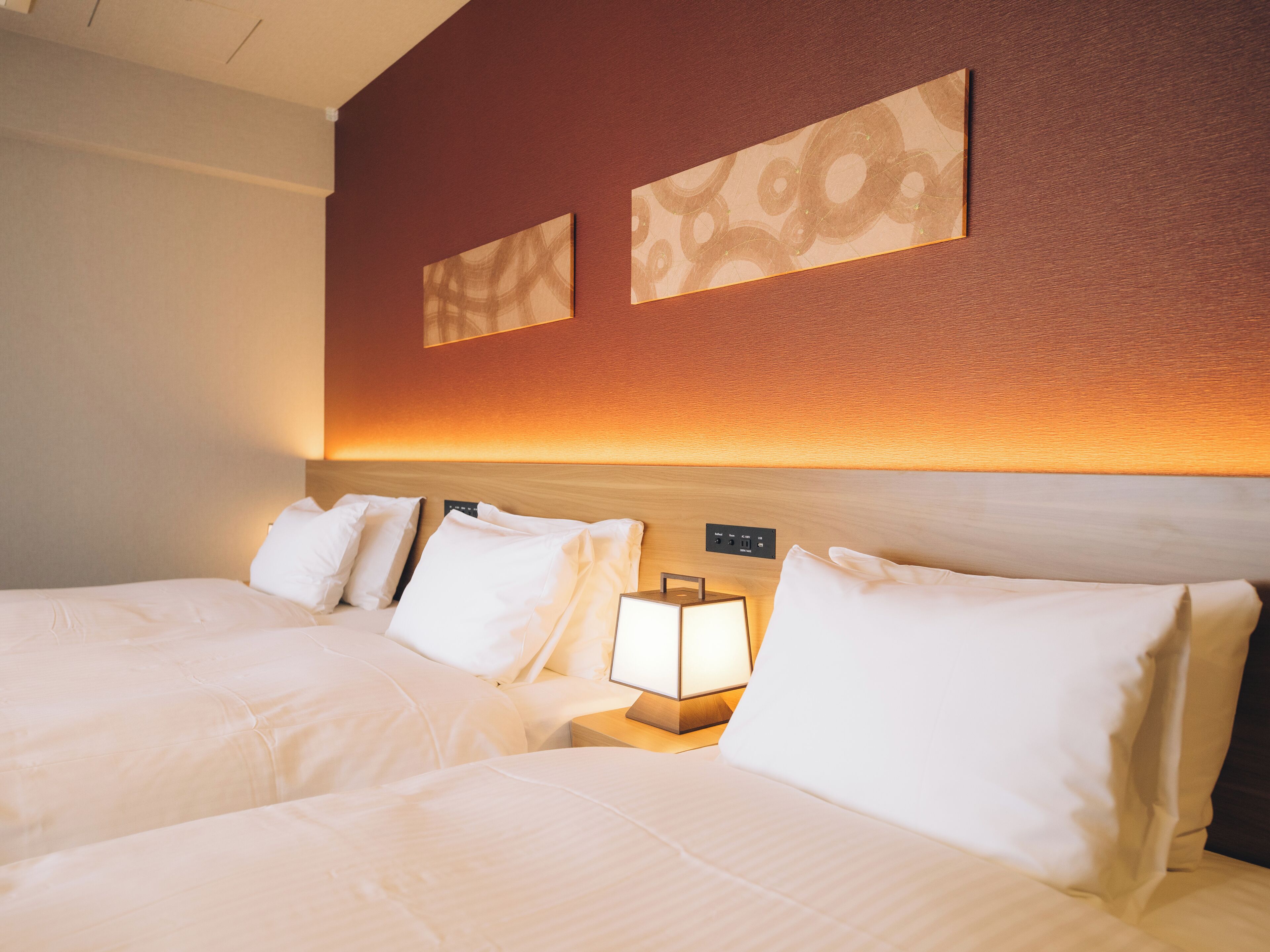 A hotel room with two beds, a nightstand with a lamp, and a wall-mounted control panel.
