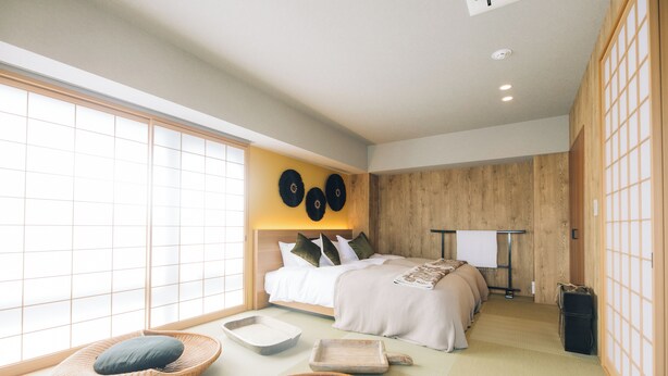 Un dormitorio de estilo japonés tradicional con suelo de tatami, una cama baja con cabecera de madera y puertas correderas shoji en las ventanas.