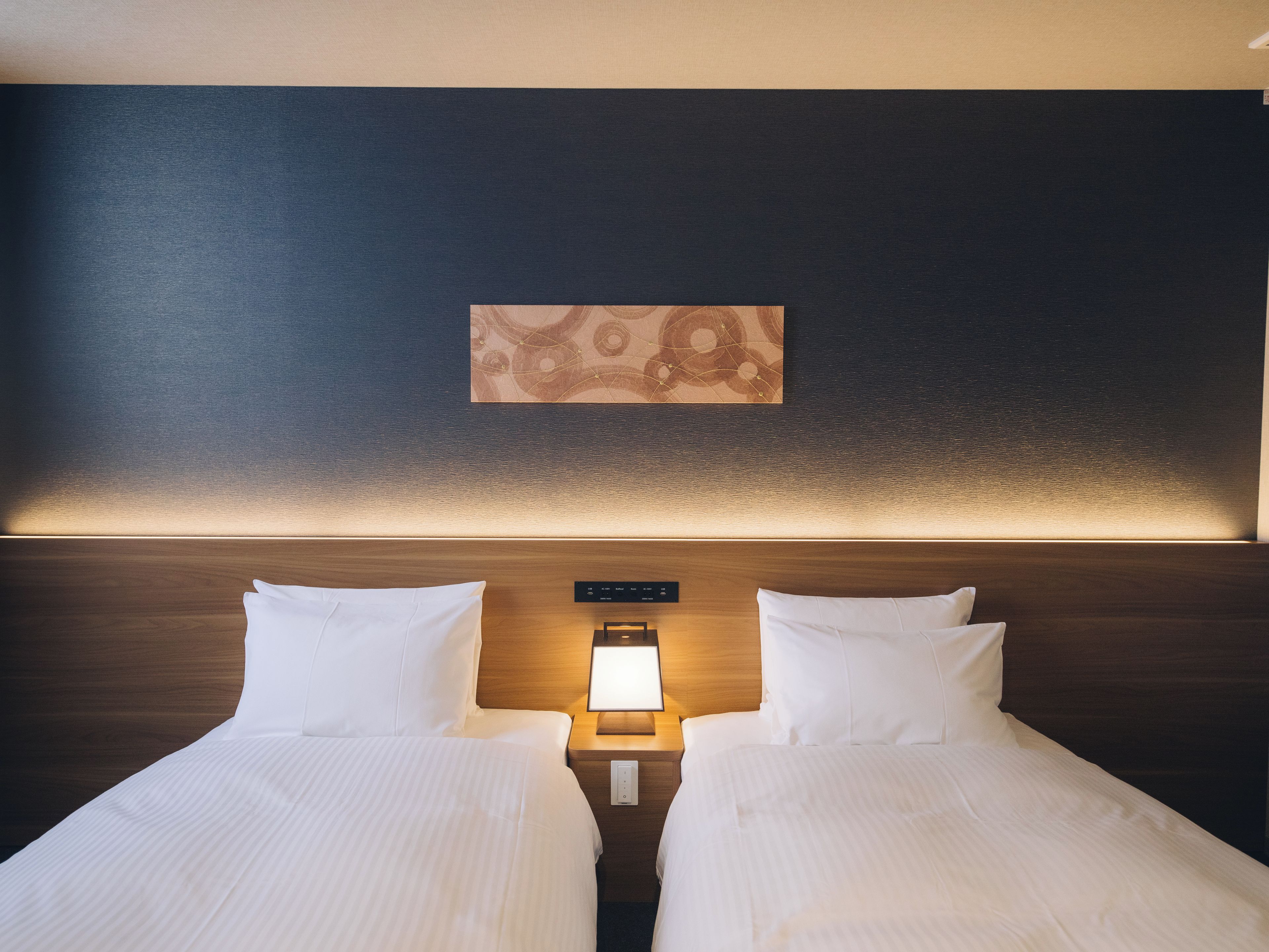 A hotel room with two beds, a nightstand with a lamp, and a wall-mounted artwork.