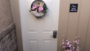 Een witte deur met een decoratieve bloem, een bord met de tekst 'Pas op uw neus', en een vaas met roze bloemen op de grond.