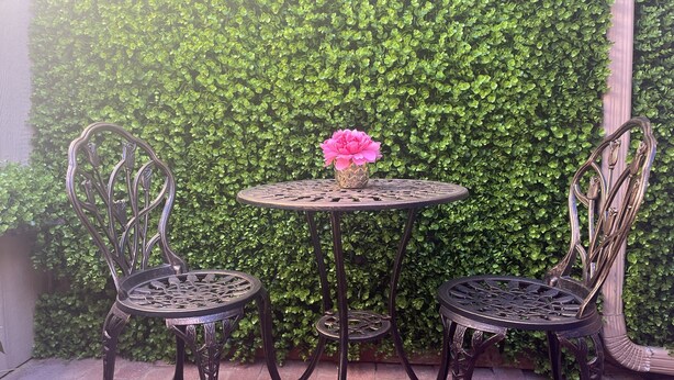 Een terras met twee metalen stoelen en een ronde tafel, met een groene, bladerrijke muur op de achtergrond.