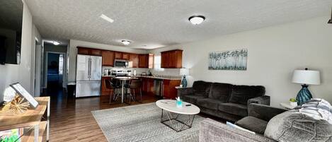 A modern living room with a sofa, coffee table, and a rug. There is a kitchen in the background with wooden cabinets and a refrigerator.