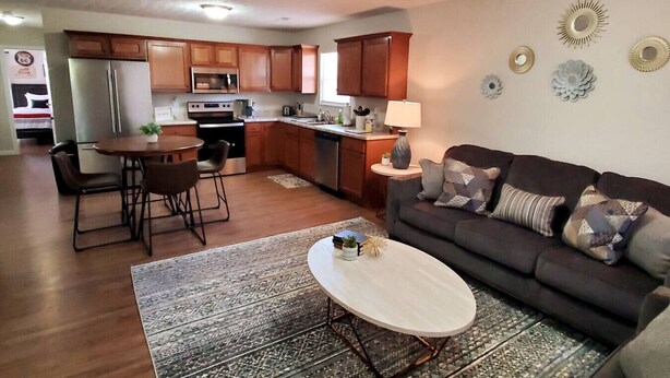 Una cocina moderna con armarios de madera, un comedor con mesa y sillas redondas, una sala de estar con sofá y cojines decorativos, y una mesita auxiliar con lámpara.