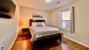 Un dormitorio con cama, mesita de noche, televisor montado en la pared y ventana con cortinas.