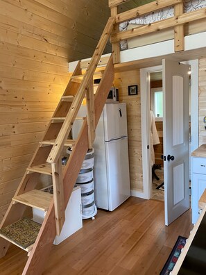 Interior de madera con una escalera de madera, un refrigerador blanco y una puerta que da a otra habitación.