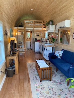 Interior acogedor de una cabaña con paredes de madera, un sofá azul, una mesa de madera y una zona de cocina con refrigerador y lavaplatos.