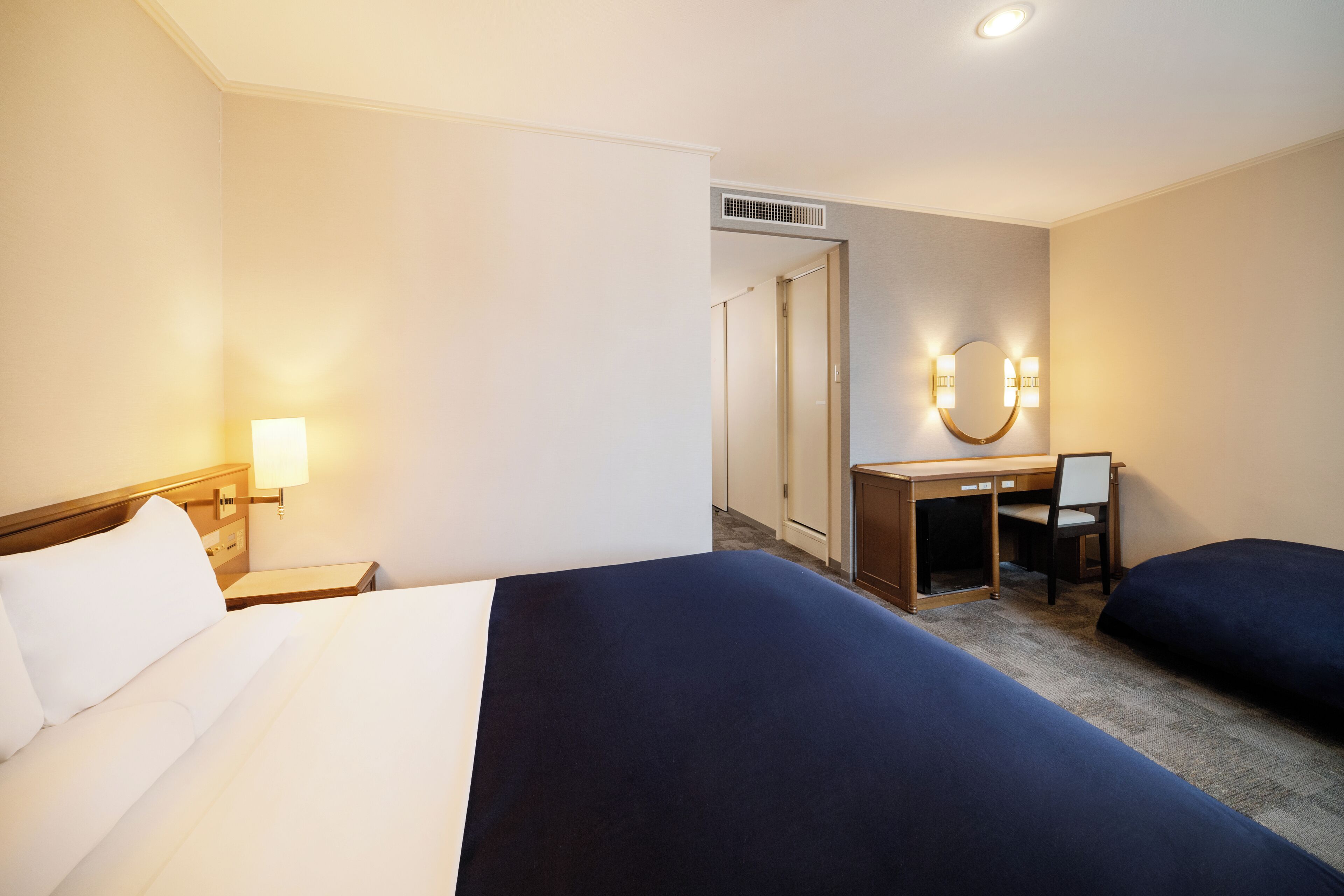 A hotel room with two beds, a desk, a chair, a mirror, and a lamp.