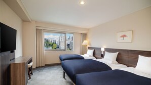 A hotel room with two beds, a desk, a TV, a window with a city view, and a lamp.