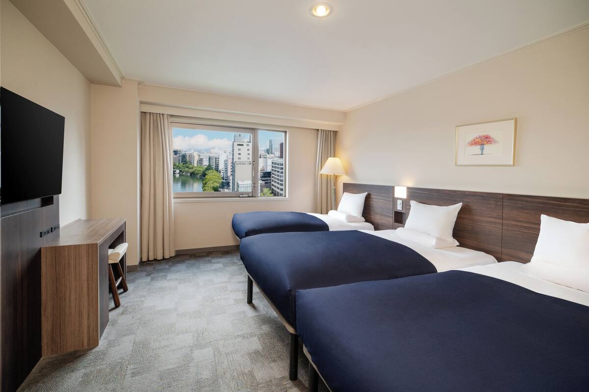 A hotel room with two beds, a desk, a TV, a window with a city view, and a lamp.