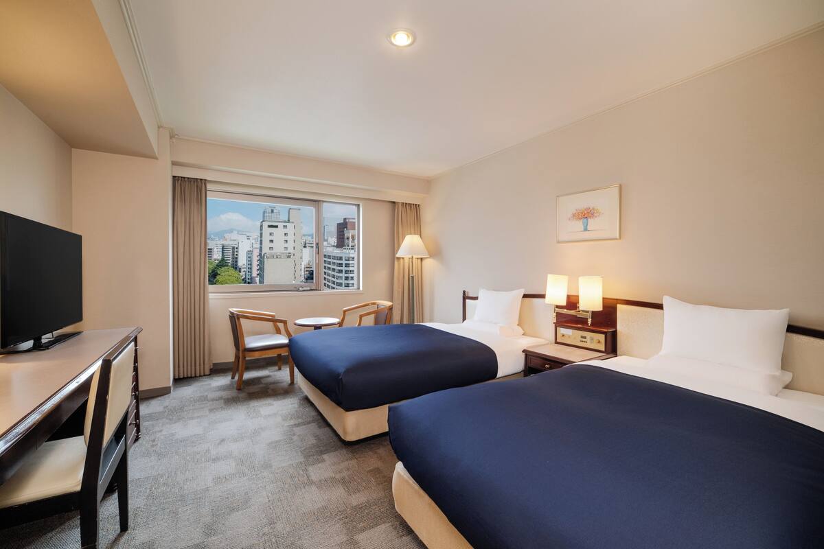 A hotel room with two beds, a desk with a TV, a chair, a lamp, and a window with a city view.