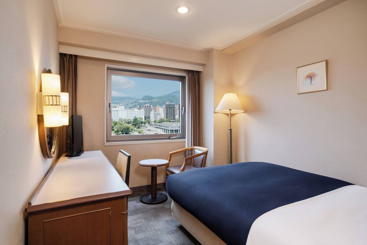 A hotel room with a bed, a desk, a chair, a window with a view of buildings and mountains, and two wall-mounted lamps.