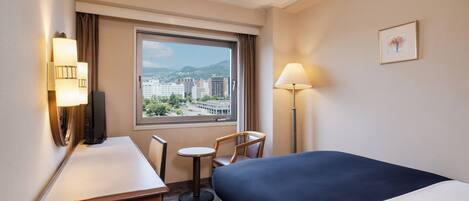 Habitación de hotel con cama, escritorio, silla, ventana con vistas a edificios y montañas, y dos lámparas de pared.