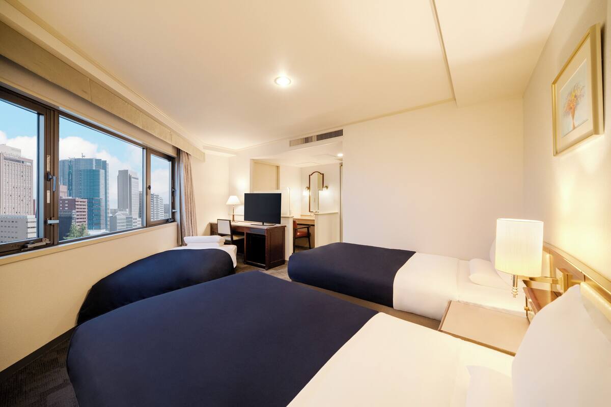 A hotel room with two beds, a desk, a television, and a large window offering a city view.