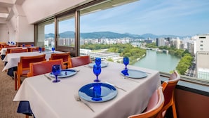 A dining area with tables set for two, large windows offering a view of a river and cityscape.