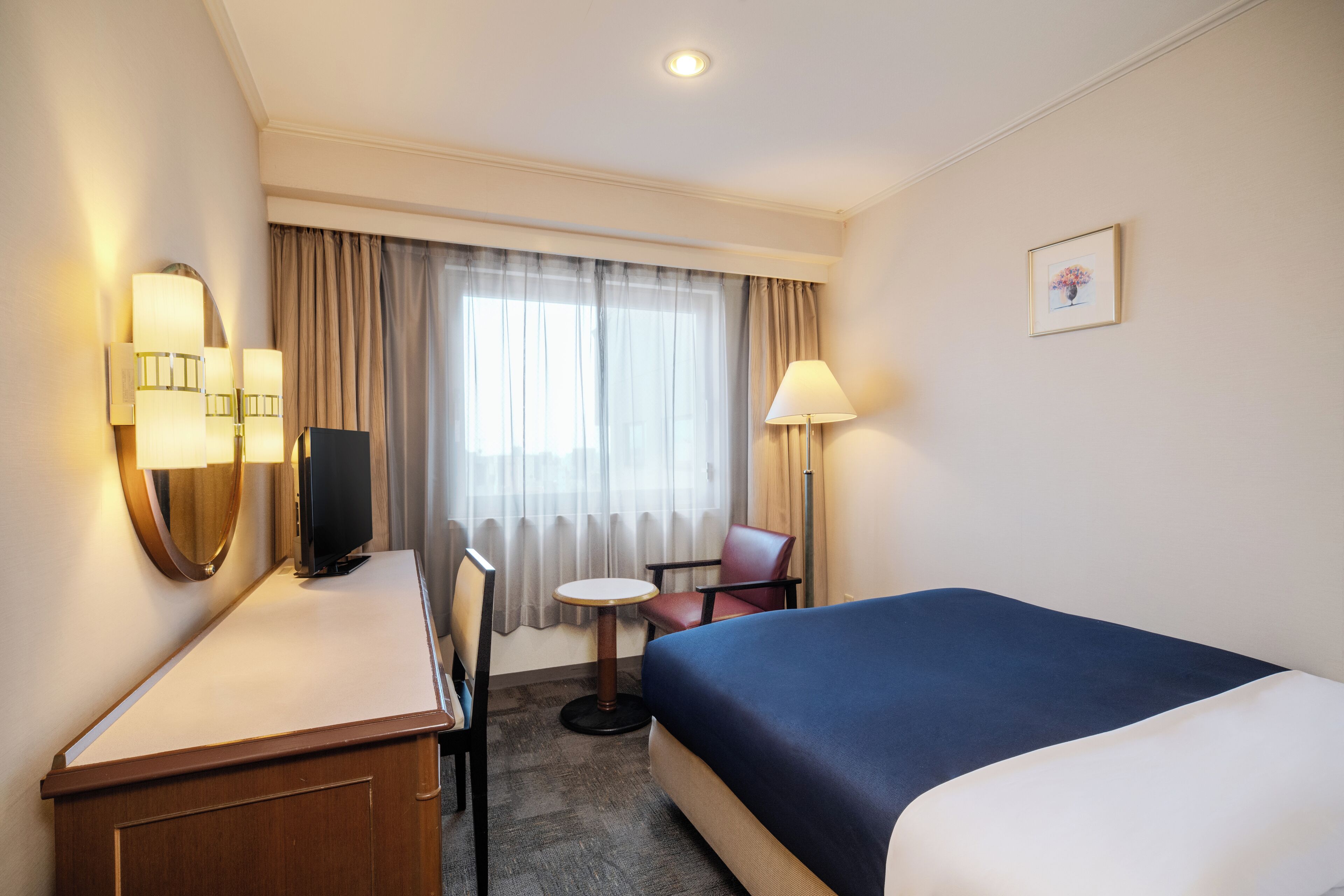A hotel room with a bed, a desk, a chair, a television, a lamp, and a window with curtains.