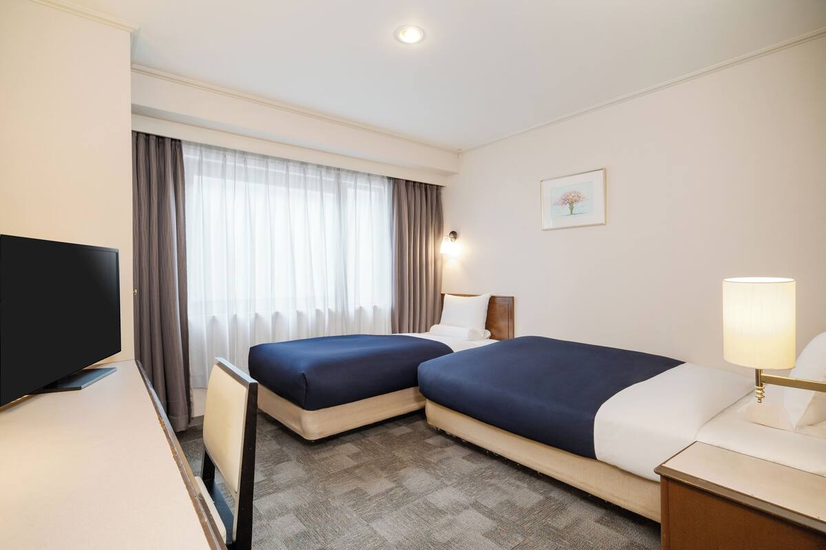 A hotel room with two beds, a desk, a TV, and a window with curtains.