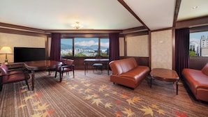 A hotel room with a large window, a flat-screen TV, a dining table with chairs, a sofa, and a small table.