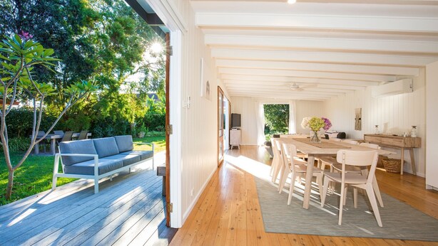 A modern open-plan living space with a wooden deck, a sofa, a dining area with chairs, and a kitchen visible through an open doorway. - Beachwood - Firepit - Pet Friendly - 2 Mins Walk to the Beach (Callala Beach)