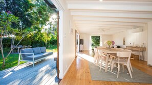 Un espacio habitable moderno y abierto con una terraza de madera, un sofá, una zona de comedor con sillas y una cocina visible a través de un vano abierto.
