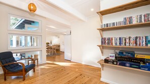A modern living room with a wooden floor, a blue armchair, a bookshelf filled with books, and a dining area in the background. - Beachwood - Firepit - Pet Friendly - 2 Mins Walk to the Beach (Callala Beach)
