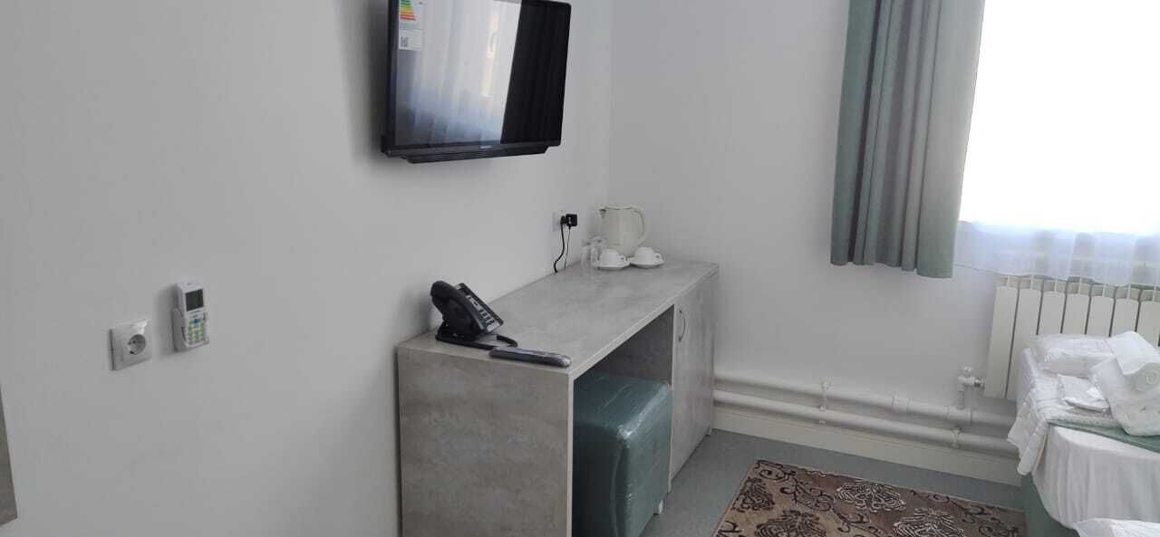 Habitación de hotel con televisión de pantalla plana en la pared, un escritorio pequeño con un teléfono, una tetera y una taza encima, un radiador y una ventana con cortinas.