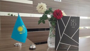 A reception desk with a blue flag, a vase with flowers, and a black sign with 'SULO Aksay' written on it. - Sulo Aksay Hotel (Aksay)