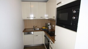 Uma cozinha compacta com armários brancos, uma pia de aço inoxidável, um forno preto e uma cafeteira na bancada.