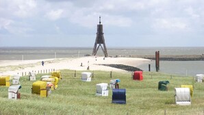 Uma área costeira com uma estrutura imponente que lembra um farol ou torre, cercada por terreno gramado e uma praia de areia.